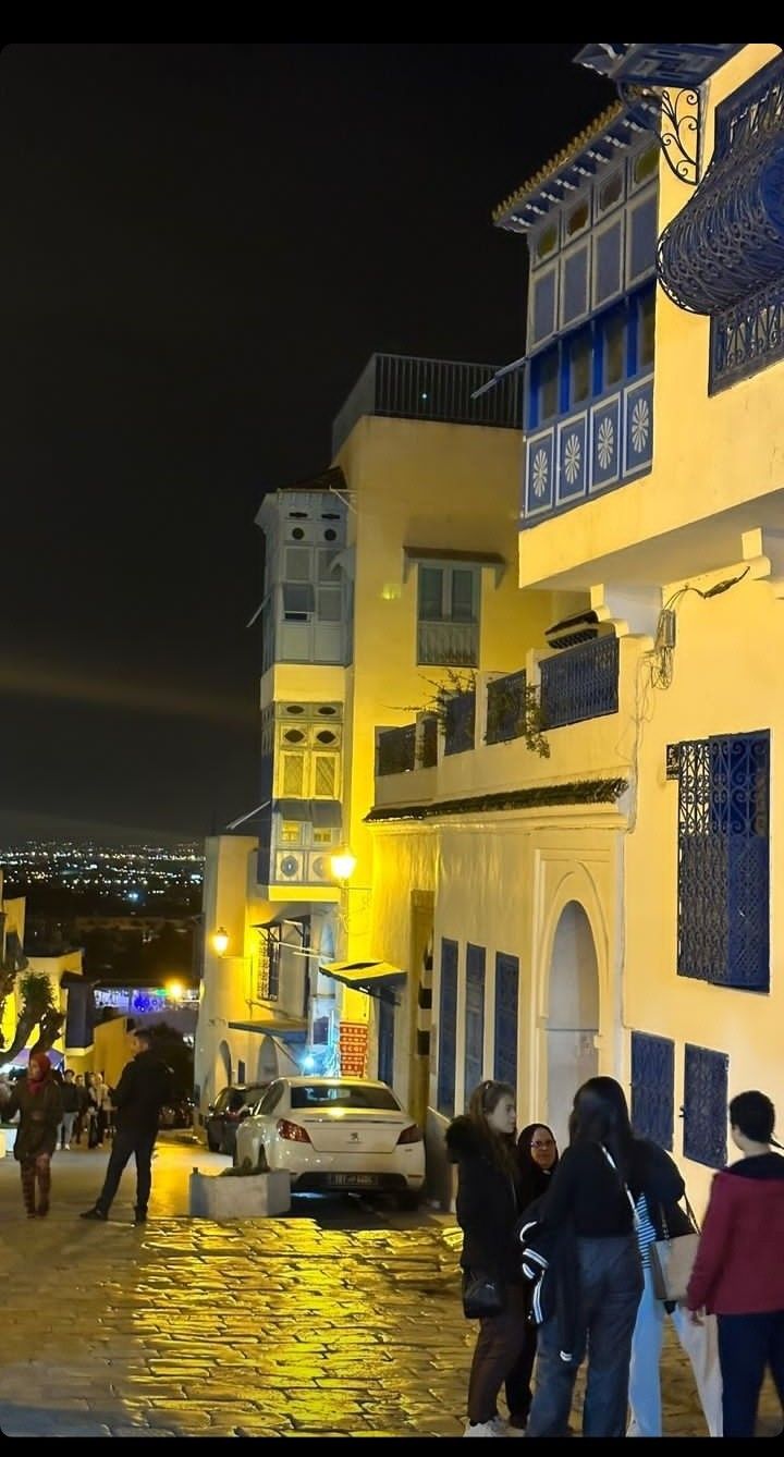 several people are walking down the street in front of some buildings at night with lights on