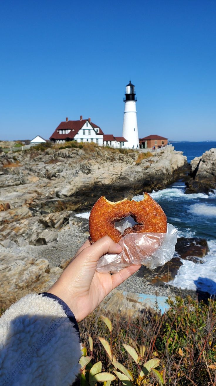 a person holding up a donut in front of the ocean with a lighthouse in the background