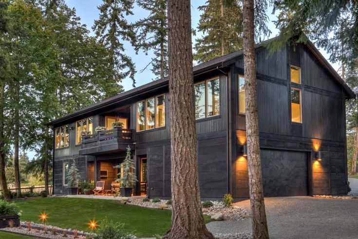 a large black house surrounded by trees in the evening time with lights on it's windows