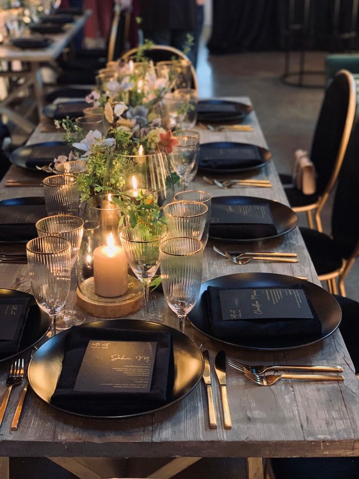 a long table set with black plates and silverware, candles and flowers in vases