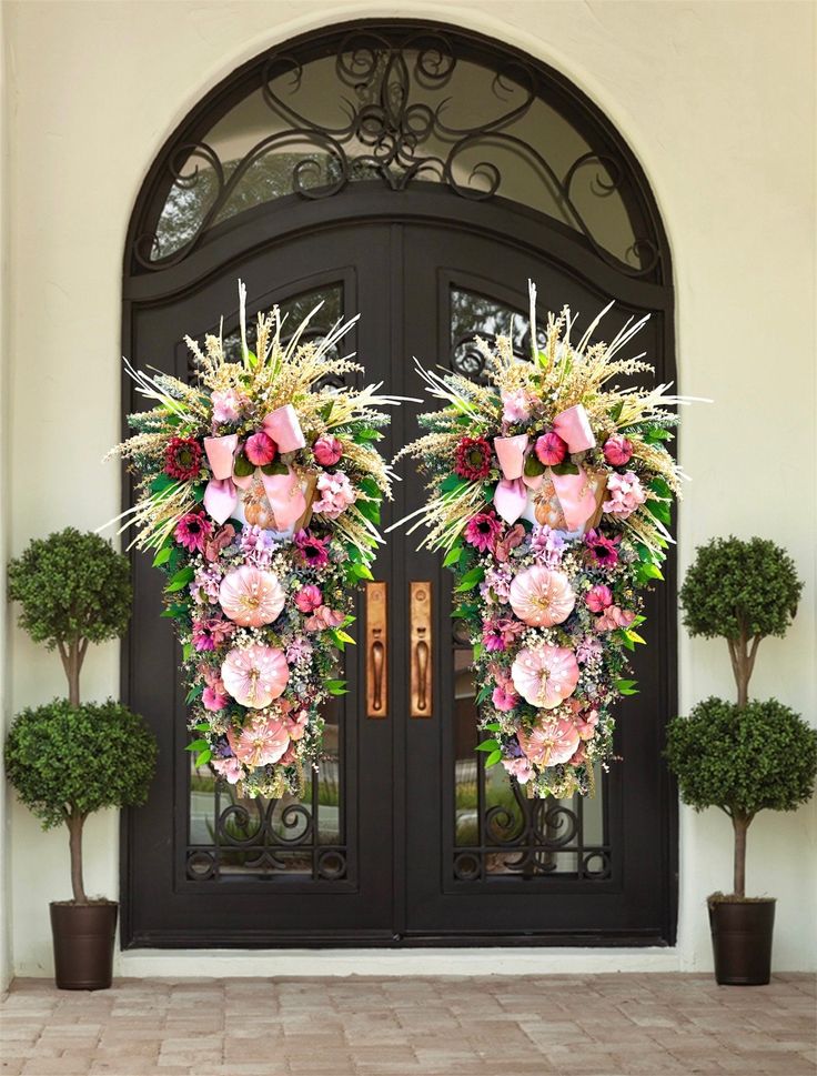 two floral wreaths on the front door of a house with potted plants and trees