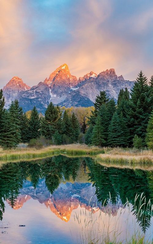 a mountain range is reflected in the still water of a lake at sunset with trees and grass around it