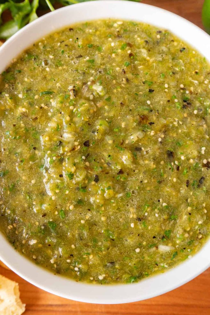 a white bowl filled with green salsa on top of a wooden table next to bread