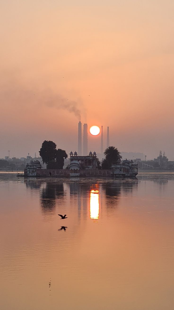 the sun is setting over a lake with birds flying around it and buildings in the background