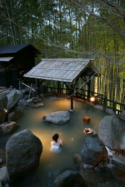 a woman in a hot tub surrounded by rocks and trees at night with the lights on