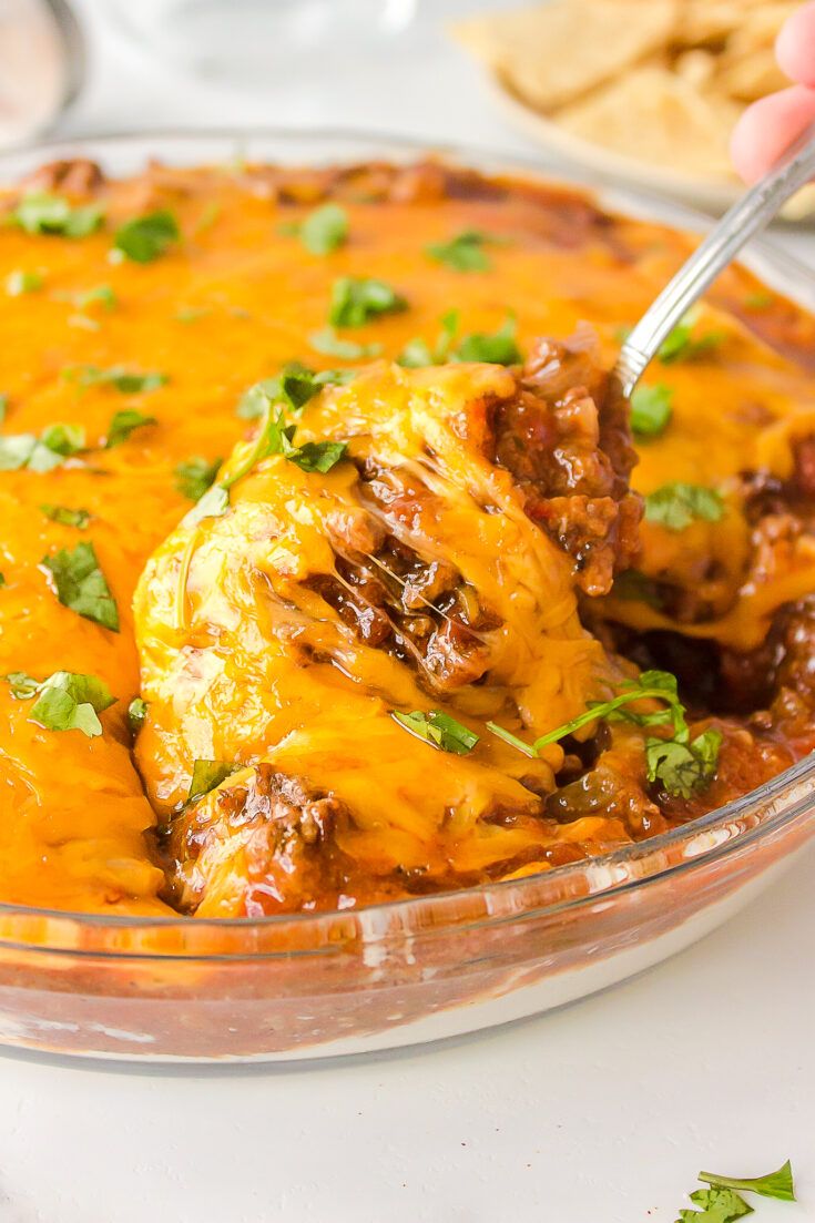 a hand holding a spoon over a casserole dish filled with meat and cheese