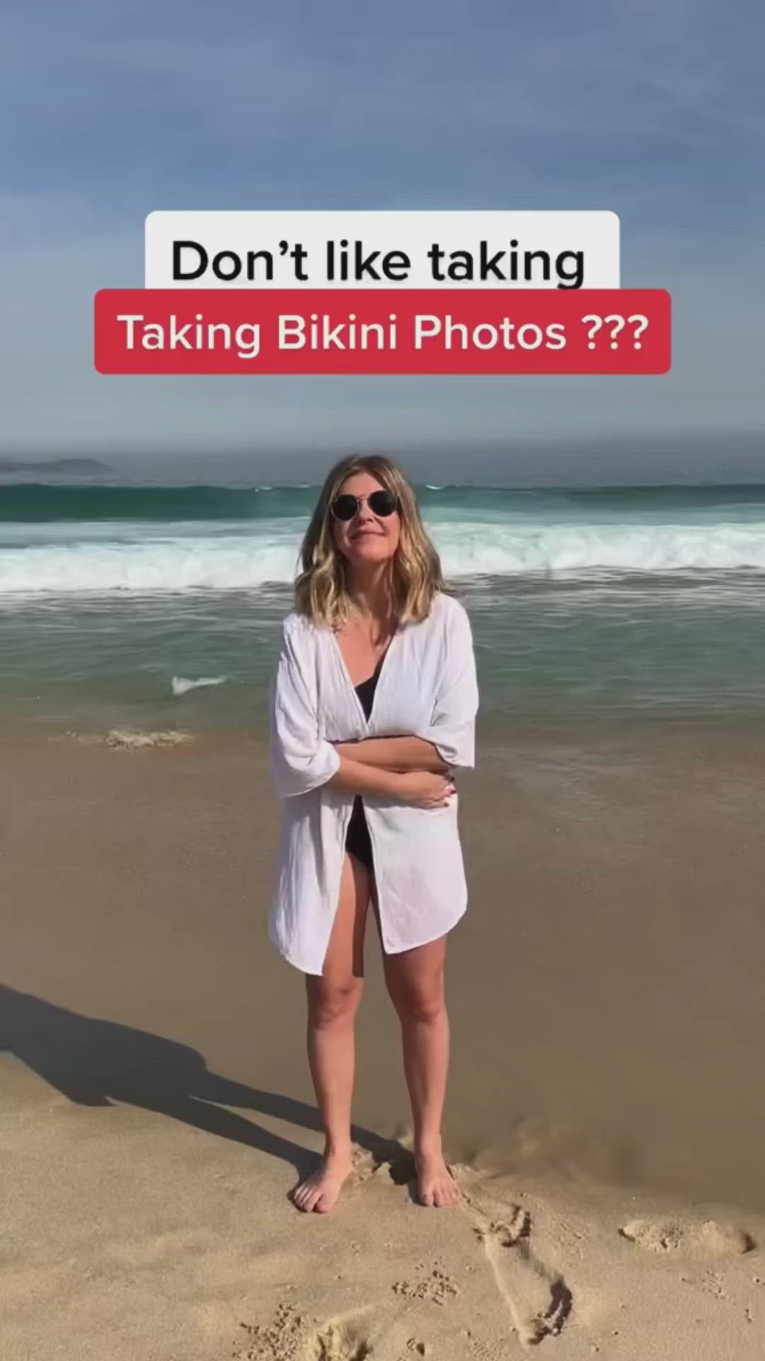 This may contain: a woman standing on top of a sandy beach next to the ocean with her arms crossed