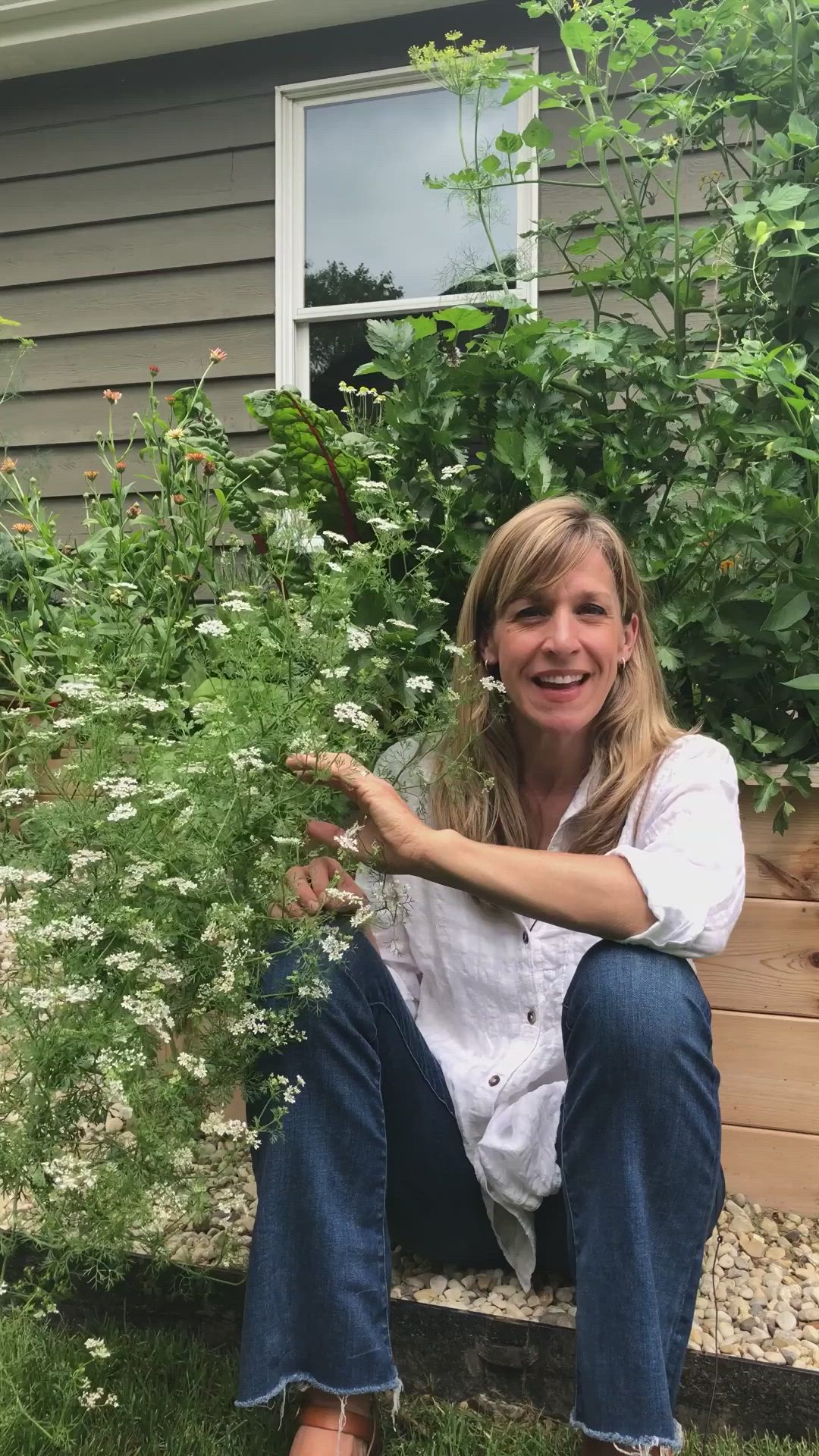 This may contain: a woman sitting on a bench in front of a house with flowers and plants around her