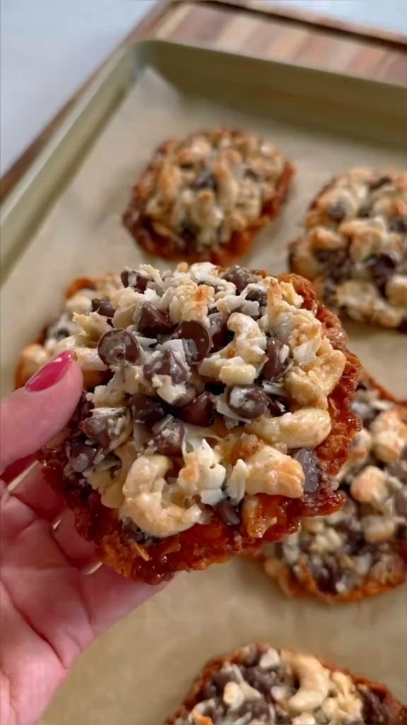 This contains: A batch of golden-brown magic cookies with cashews, coconut flakes, and chocolate chips on a baking sheet.