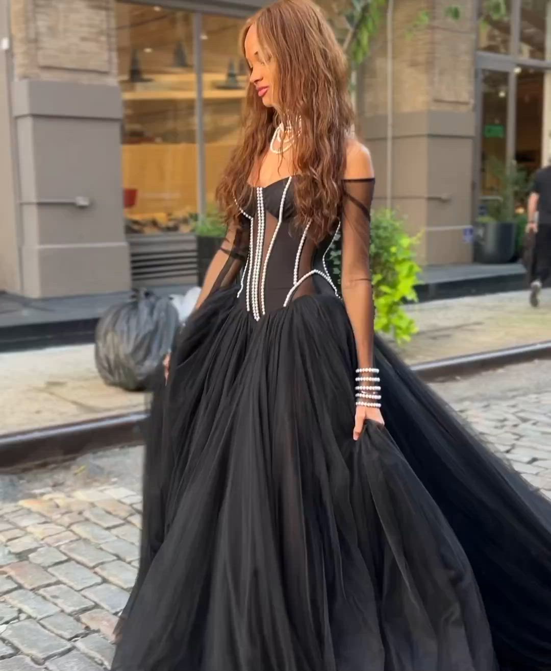 Embrace the drama in this black ball gown extravaganza. A symphony of elegance unfolds as a dramatic, pearl-studded boned sheer corset takes center stage, adorned with opulence. The corset's intricate design is paired with an opulent black tulle skirt, creating an ethereal dance of textures.