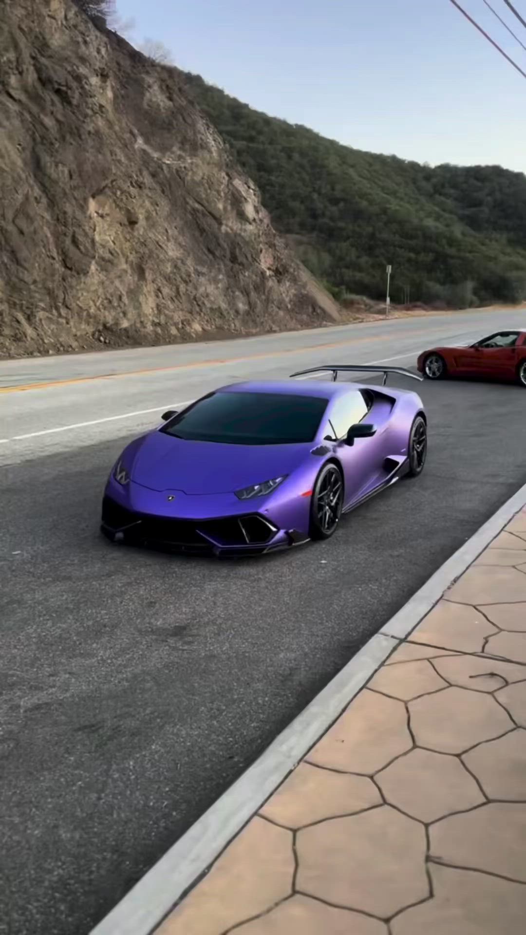 This may contain: two cars are parked on the side of the road near some hills and mountains, one is purple