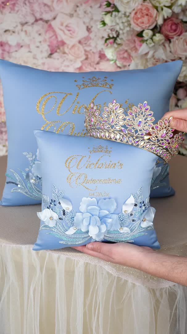 This may contain: two blue pillows with gold lettering on them and flowers in the background, one being held by a woman's hand