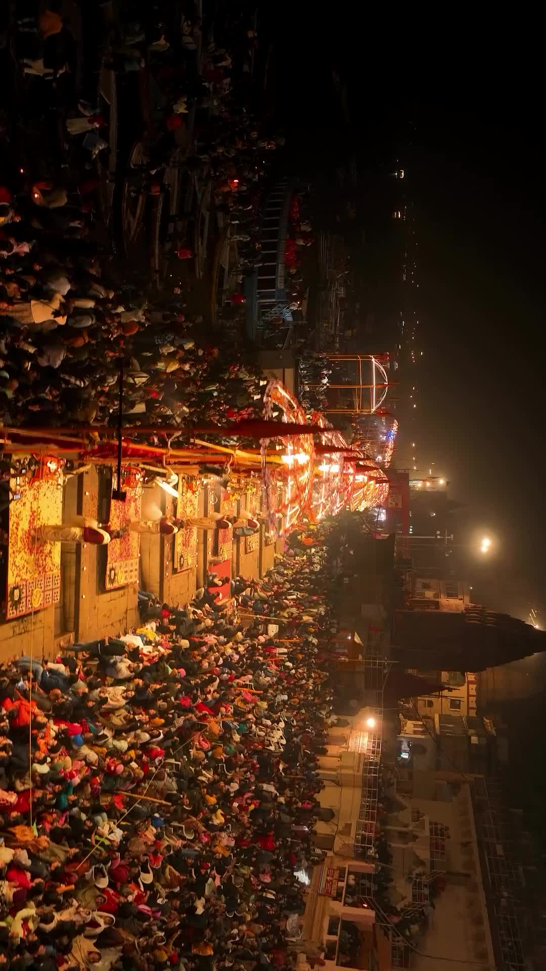 This may contain: a large crowd of people standing on the side of a river at night with lights and umbrellas