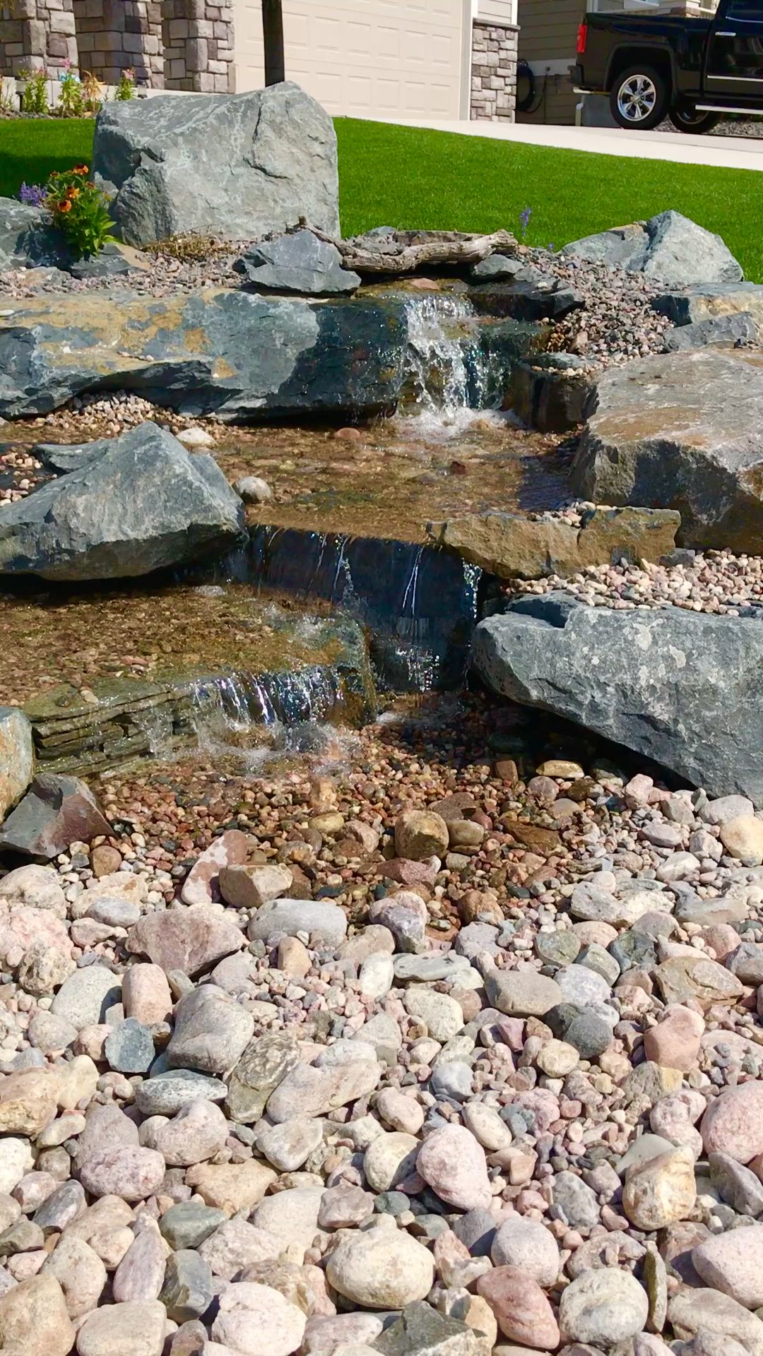 This may contain: a rock garden with rocks and water flowing from it to the ground, in front of a house