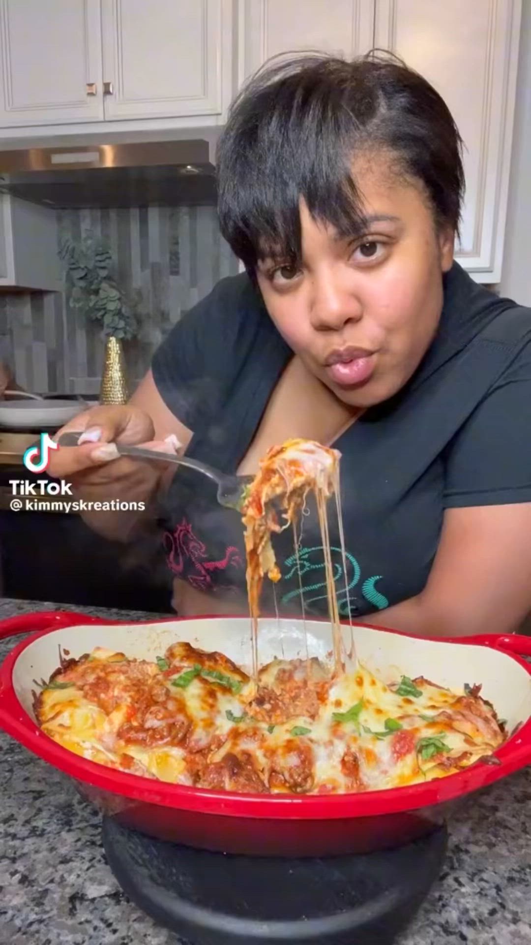 This may contain: a woman is eating spaghetti from a red casserole dish on the kitchen counter