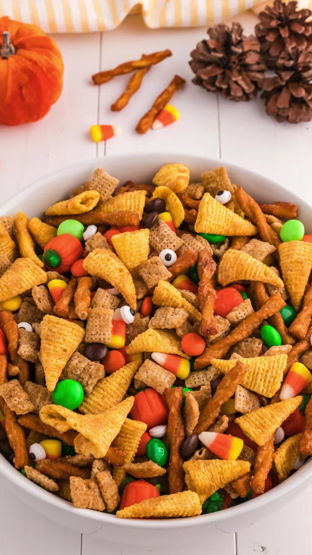 This may contain: a white bowl filled with halloween cheetos and candy cornflakes next to pine cones