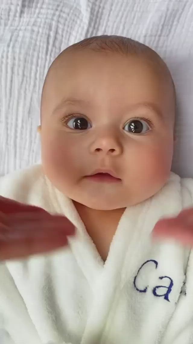 This may contain: a baby laying on top of a bed wearing a white robe and pointing at the camera