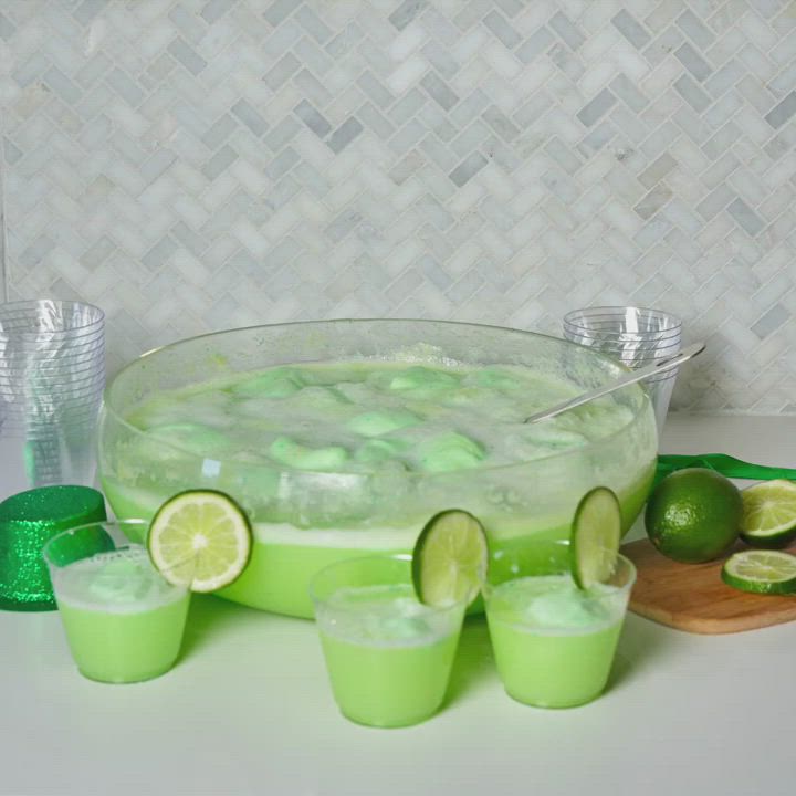 This may contain: limeade punch in a glass bowl surrounded by limes and shot glasses on a counter