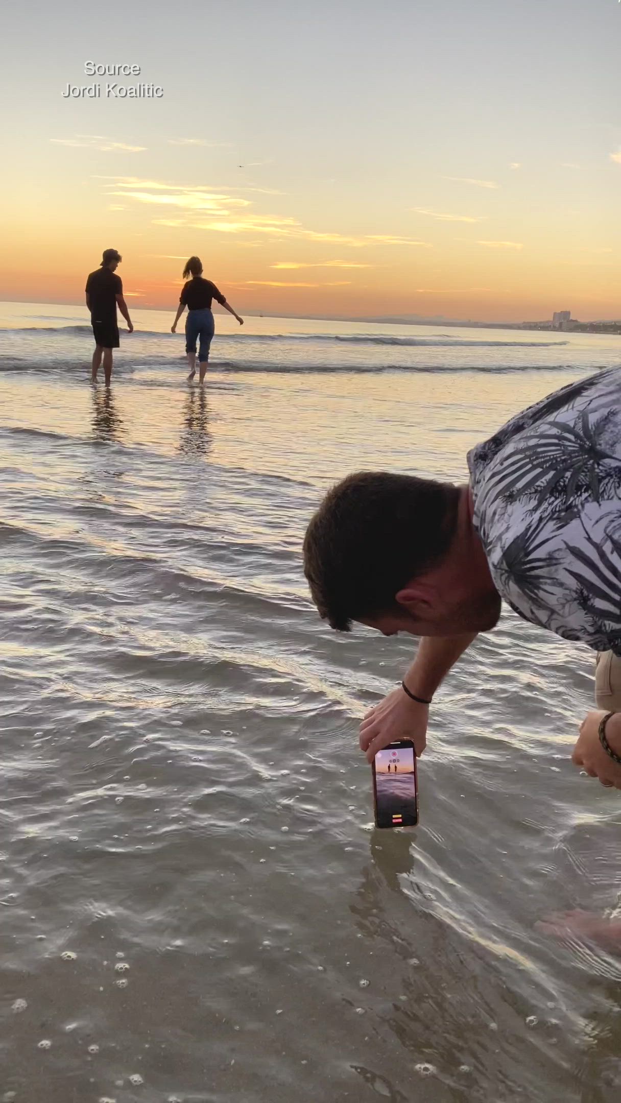 This may contain: a man is holding up his cell phone in the water while others stand on the beach