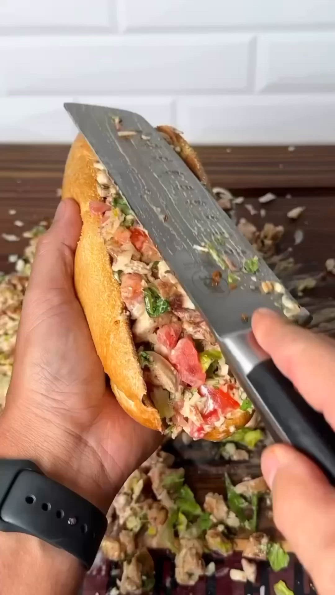 This may contain: a person holding a large knife over a sandwich with meat and vegetables on it's side