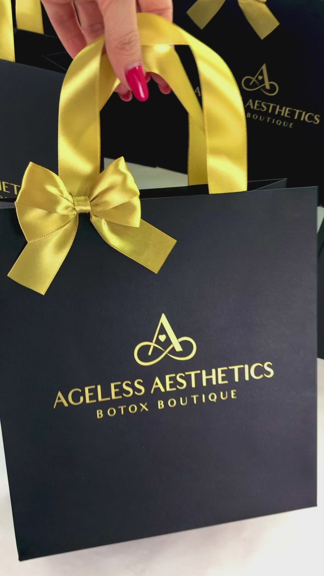 This may contain: a woman's hand holding two black shopping bags with gold ribbons on them and the logo for ageless aesthetics boston boutique