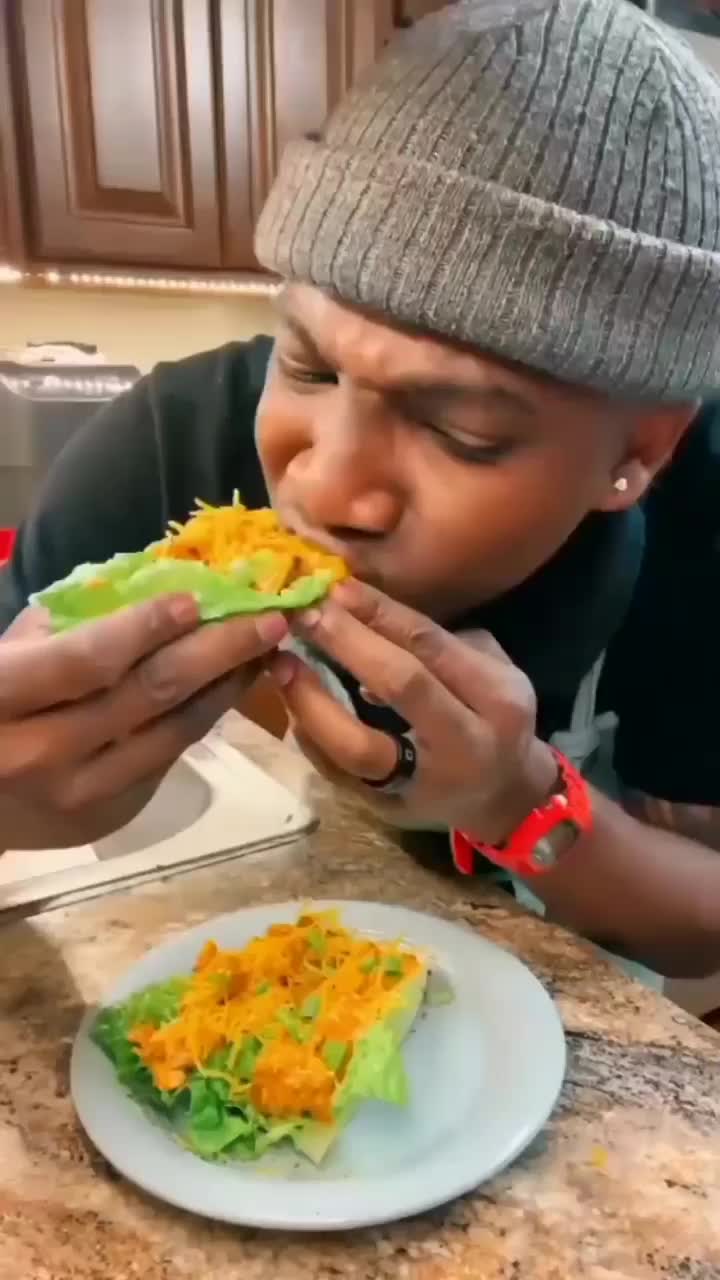 This may contain: a young man eating a sandwich with lettuce and cheese on it while wearing a beanie