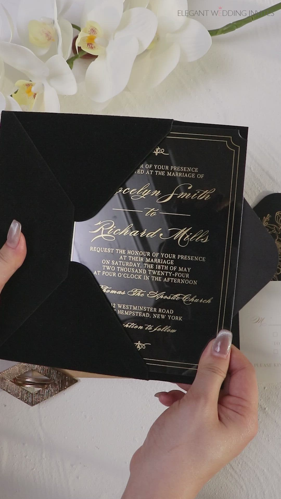 This may contain: a person holding up a black and gold wedding card with white flowers in the background