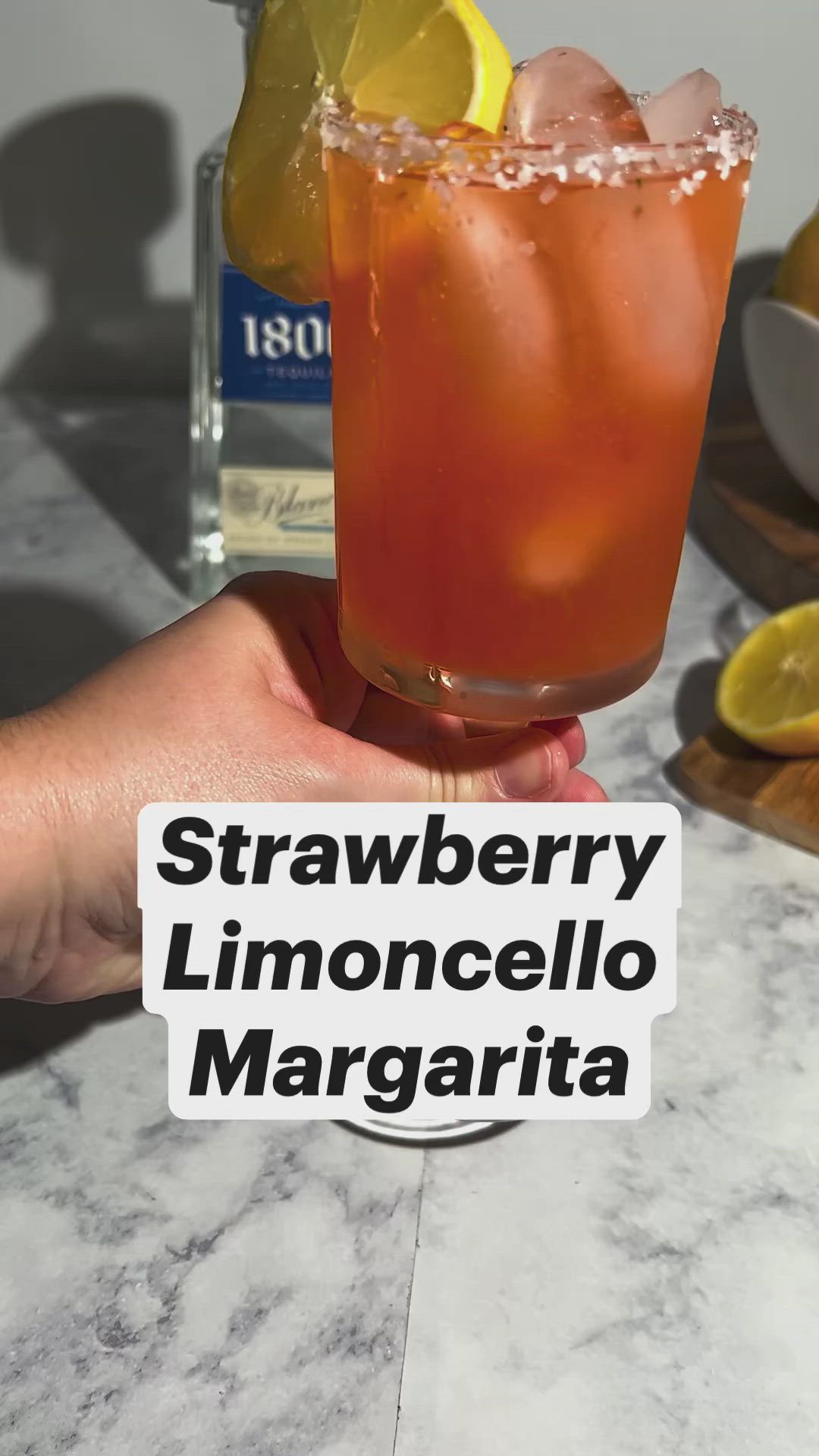 This may contain: a hand holding a drink with lemon wedges on the rim and strawberries in the background