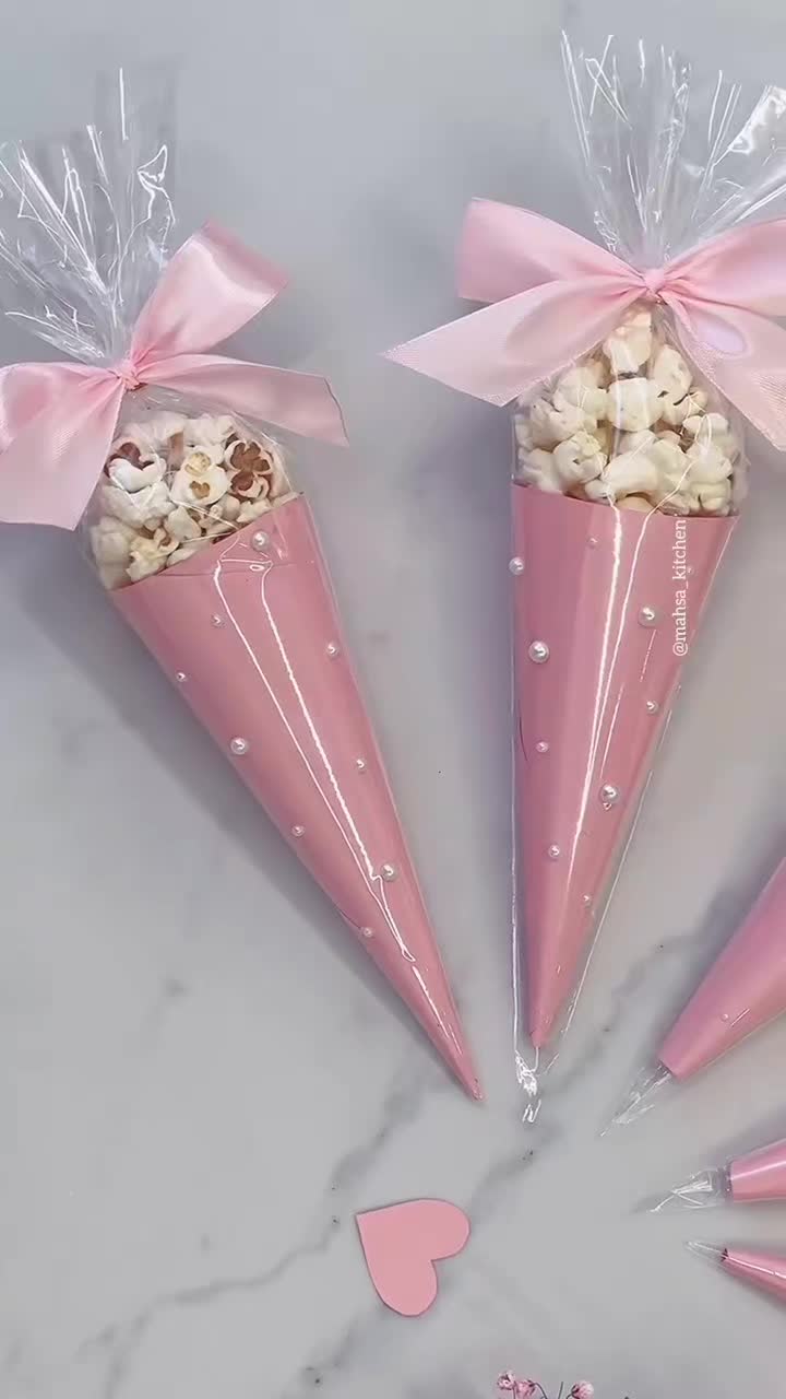 This may contain: three pink cones filled with popcorn sitting on top of a white marble counter next to hearts