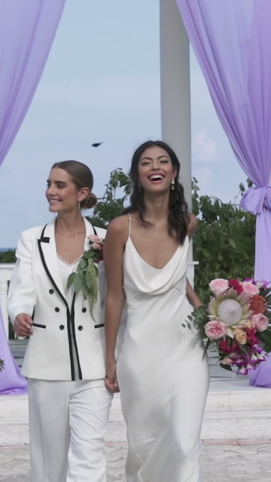 This may contain: two women in white dresses holding hands and walking down the aisle with purple drapes