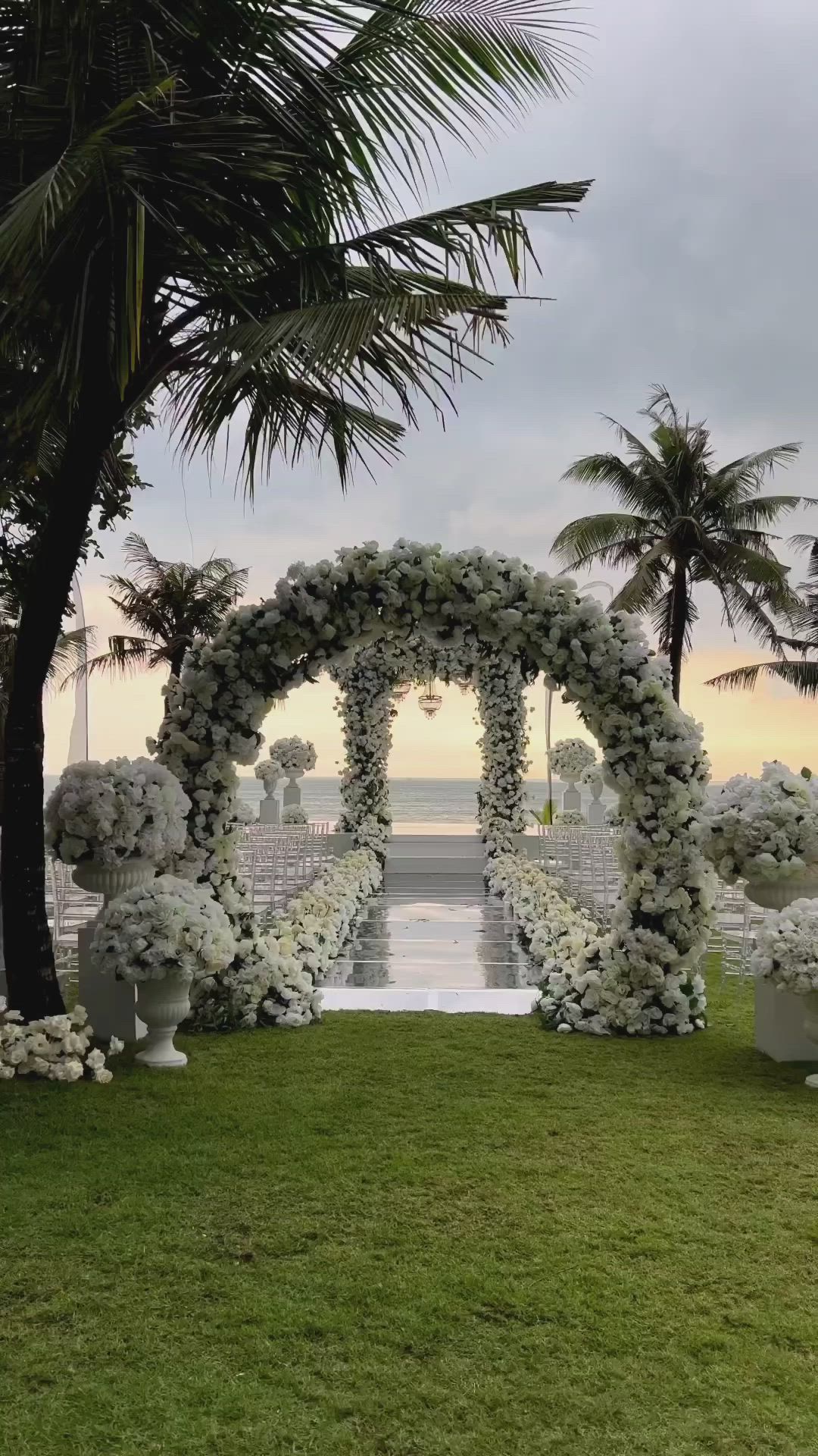 This may contain: an outdoor ceremony setup with white flowers and greenery on the grass, surrounded by palm trees