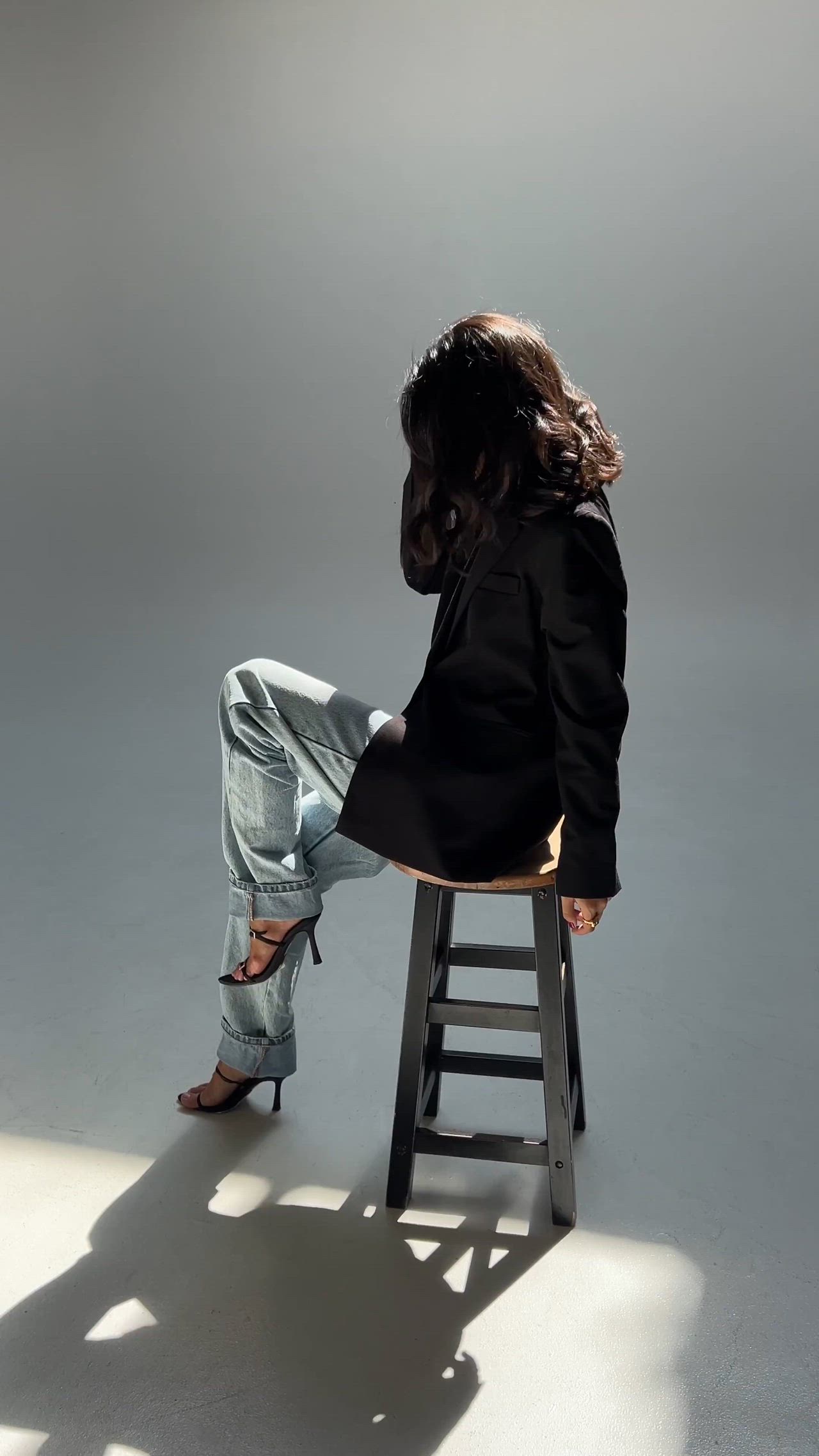 This may contain: a woman sitting on top of a wooden stool in front of a white wall with her back to the camera