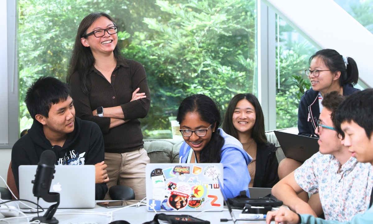 Faculty member standing and six students sitting with laptops in a conference room, with green trees visible through window. Several of them are smiling and laughing.