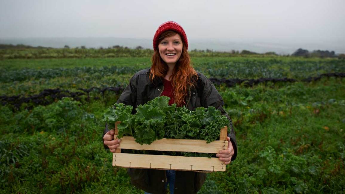 female holding a wooden crate of kale in a kale field