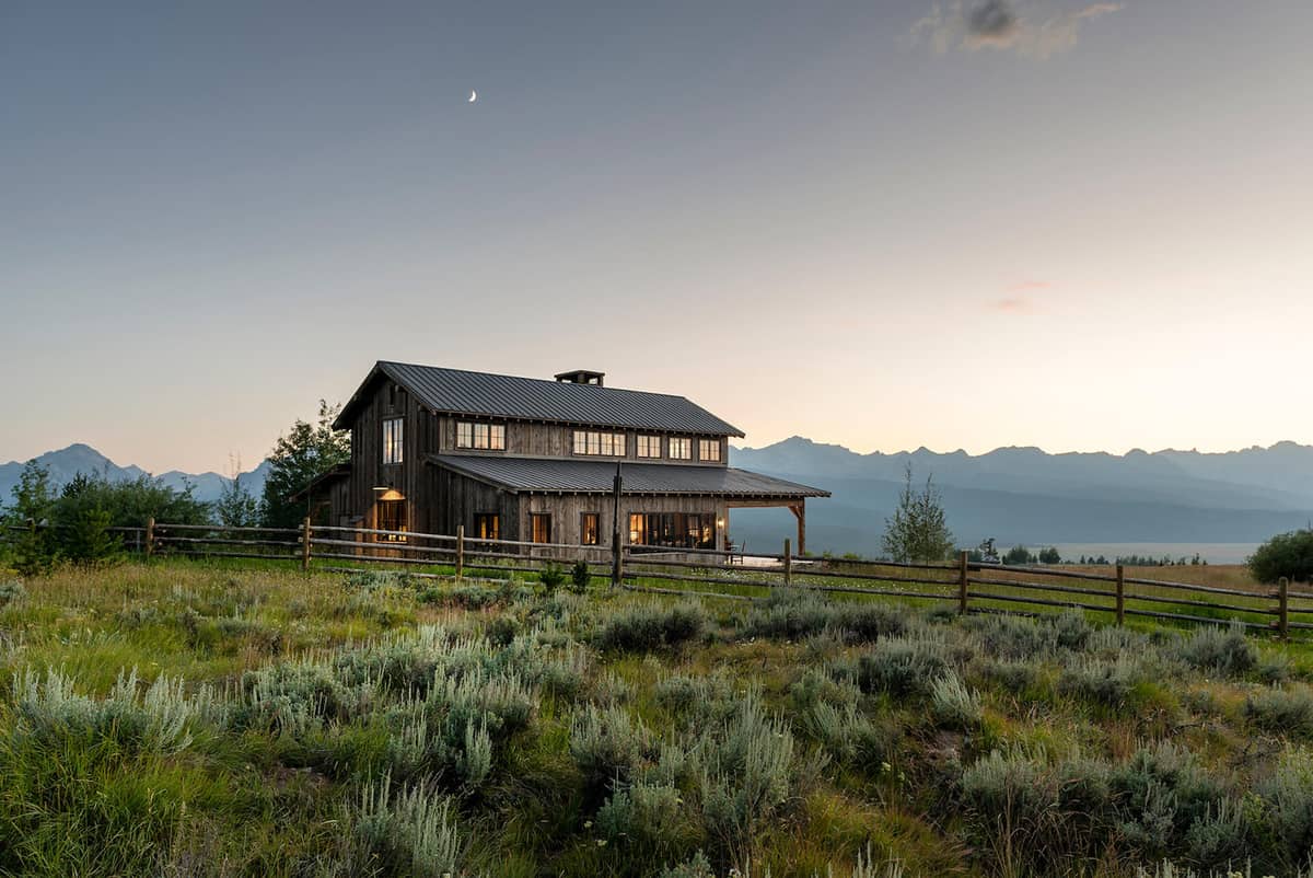 Traditional barn style house built in a grass field with a sun setting behind a mountain range in the background.