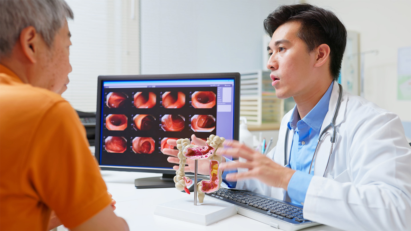 A male doctor sits next to a computer screen with diagnostic images while gesturing at a 3D model of a colon, across the desk from an older man wearing an orange shirt