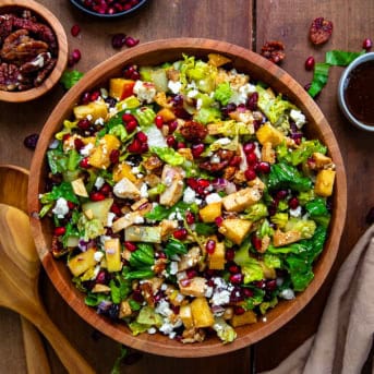 Wooden bowl of Winter Chopped Salad with lettuce, apples, pomegranate, chicken, and feta on a wooden table.