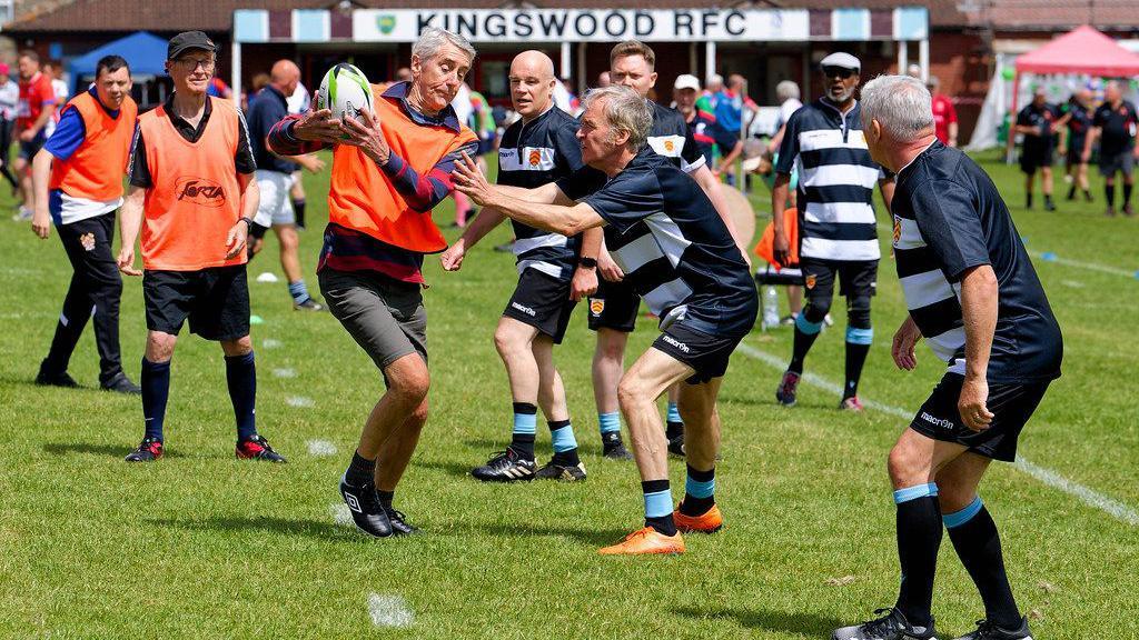 Players take part in the Kingswood Walking Rugby Championships. One team is wearing blue and white hooped tops while the other is wearing orange bibs