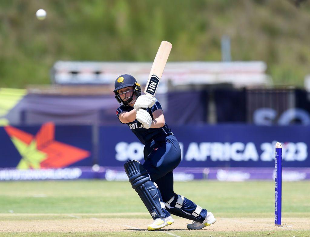 Niamh Muir in a dark blue cricket outfit plays a driving shot with her bat before setting off for a run