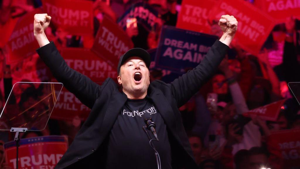 Elon Musk with his fists raised and mouth open shouting at a Trump rally. He wears a black cap, T-shirt and blazer. 