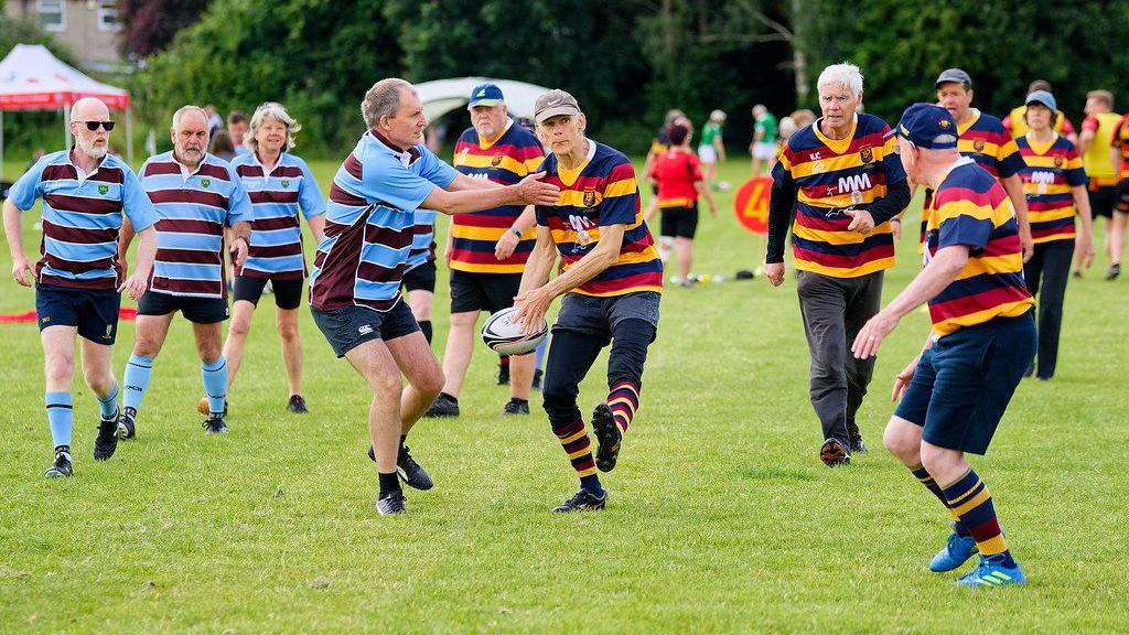 Players take part in the Kingswood Walking Rugby Festival, with one team wearing blue and brown striped tops and one in a more colourful yellow, red and blue strip. A man is being tackled as he passes the ball