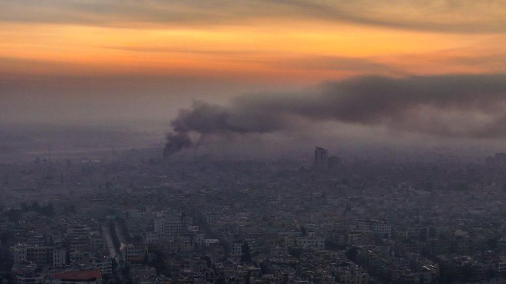 Smoke rises from an aerial view of Damascus city with sunlight in the background