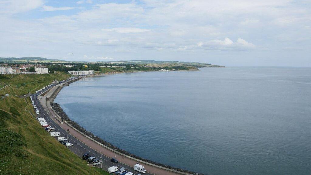 A view of the North Bay taken from adjacent to the Norbreck Hotel above Clarence Gardens. The road snakes around the sea in a half circle, lined with caravans and cars.