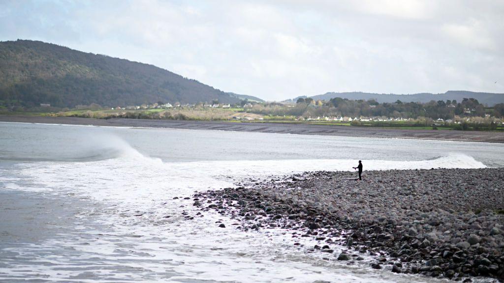 View of Pollock Weir. Waves are coming in as a surfer in the distance is getting ready. Porlock is in the background in front of a hill.