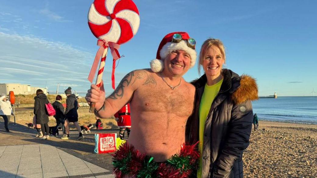 Sean and Julie Butler who is wearing a coat and jumper. Sean is wearing tinsel round his trunks and a Santa hat with goggles. 