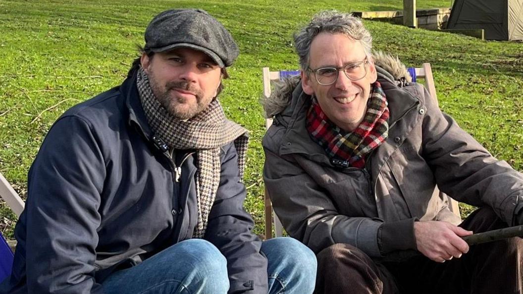 BBC Reporter Justin Dealey is sat next to Robert Wilfort on a pair of deck chairs. Both are wearing coats and scarfs. Robert is holding a fishing rod.