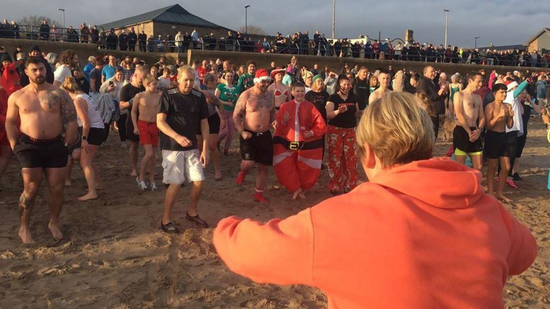 Dozens of people on the sand at Berwick, many in shorts. Some are wearing Santa hats as they prepare to run into the sea. 