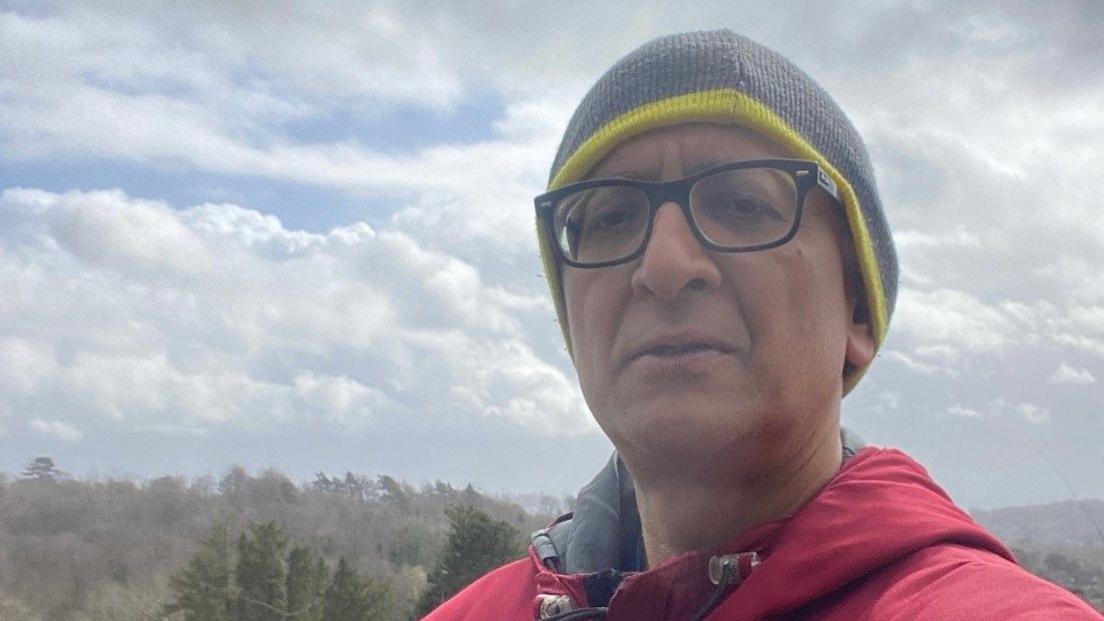 Dr Ravi Kumar - wearing black glasses, a green beanie and red jacket - taking a selfie with trees stretching into the horizon behind him