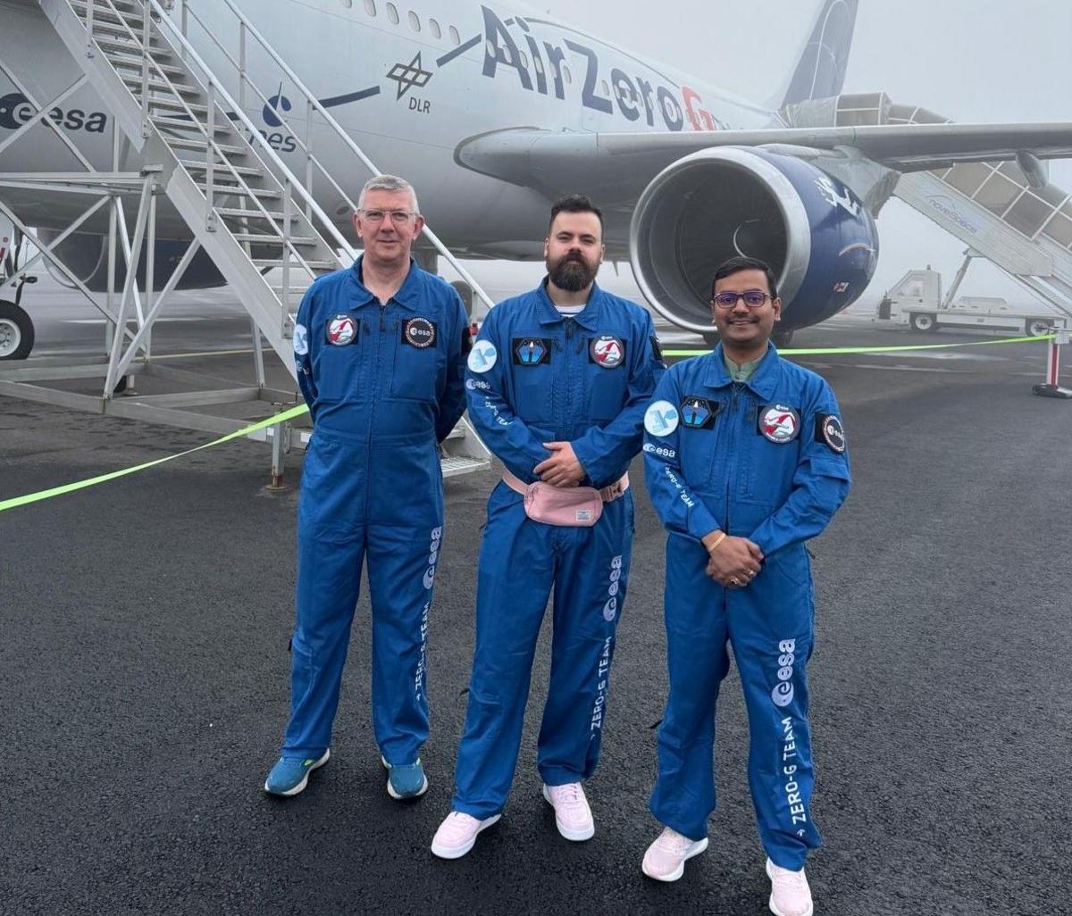 3D printing team Colin McInnes, Gilles Bailet, Satyam Bhatti, wearing blue flight suits marked with the European Space Agency Zero-G Team logo in white, stand on the tarmac in front of the Air Zero G plane.