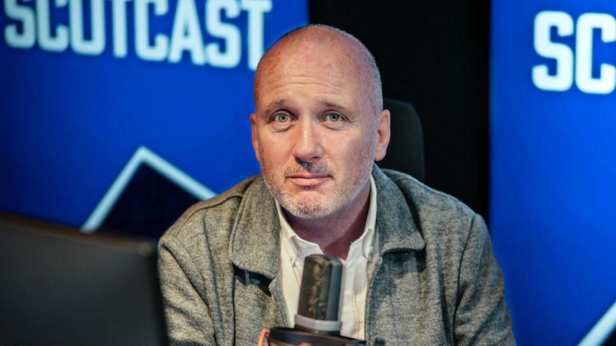 Martin Geissler sits at a microphone in a radio studio, with scotcast logo in background, wearing grey shirt with grey stubble on face.